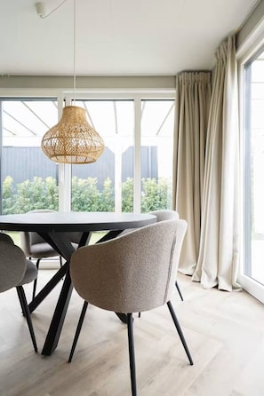 Un coin repas comprenant une table ronde, des chaises, un luminaire suspendu en fibres tressées et de grandes fenêtres avec des rideaux.