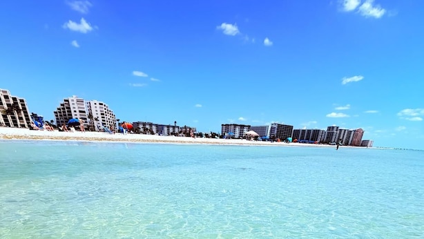 A beach with clear water, buildings in the background, and a clear sky.