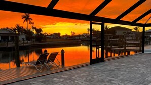A covered outdoor area with a view of a sunset over water, lounge chairs, and a dock.