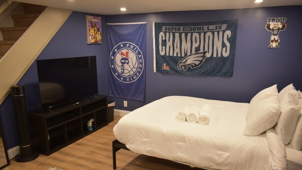 A bedroom with a bed, a TV, and sports banners on the wall. - Philly Sports Fan Suite Private Entrance + Patio Near PHL (Sharon Hill)