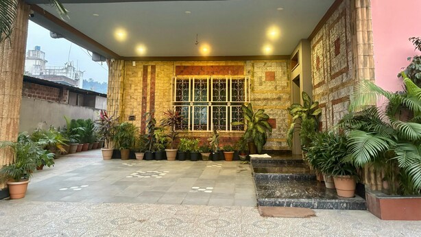 A building entrance with a tiled floor, potted plants, a window with decorative metal bars, and a stone wall with a patterned design.