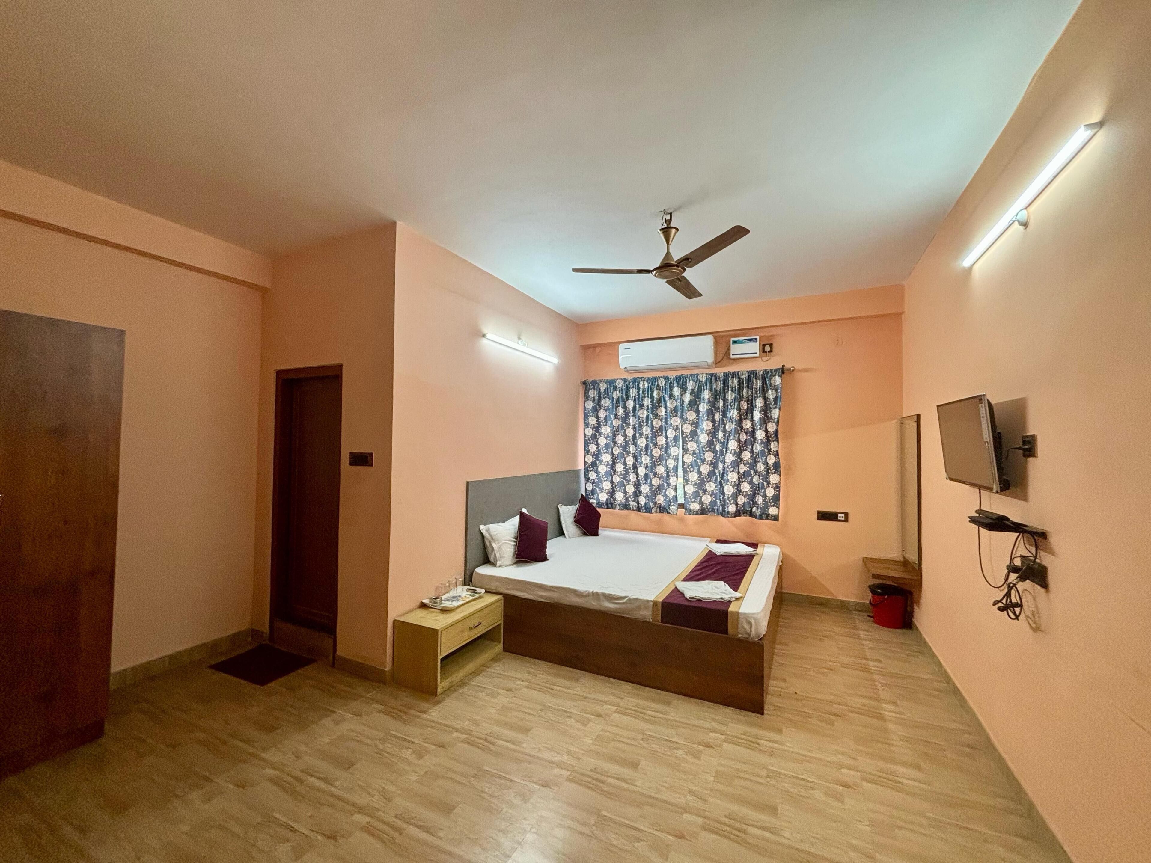 A bedroom with a bed, a ceiling fan, an air conditioning unit, a television, and a wooden floor.