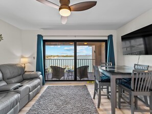Un salon moderne comprenant un canapé, une table à manger avec des chaises, un ventilateur de plafond et une vue sur l’eau, visible à travers les portes vitrées.