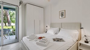Habitació d'hotel amb un llit, una tauleta de nit, un ventilador de sostre, una finestra amb vistes a vegetació i una porta que dona a zona exterior.