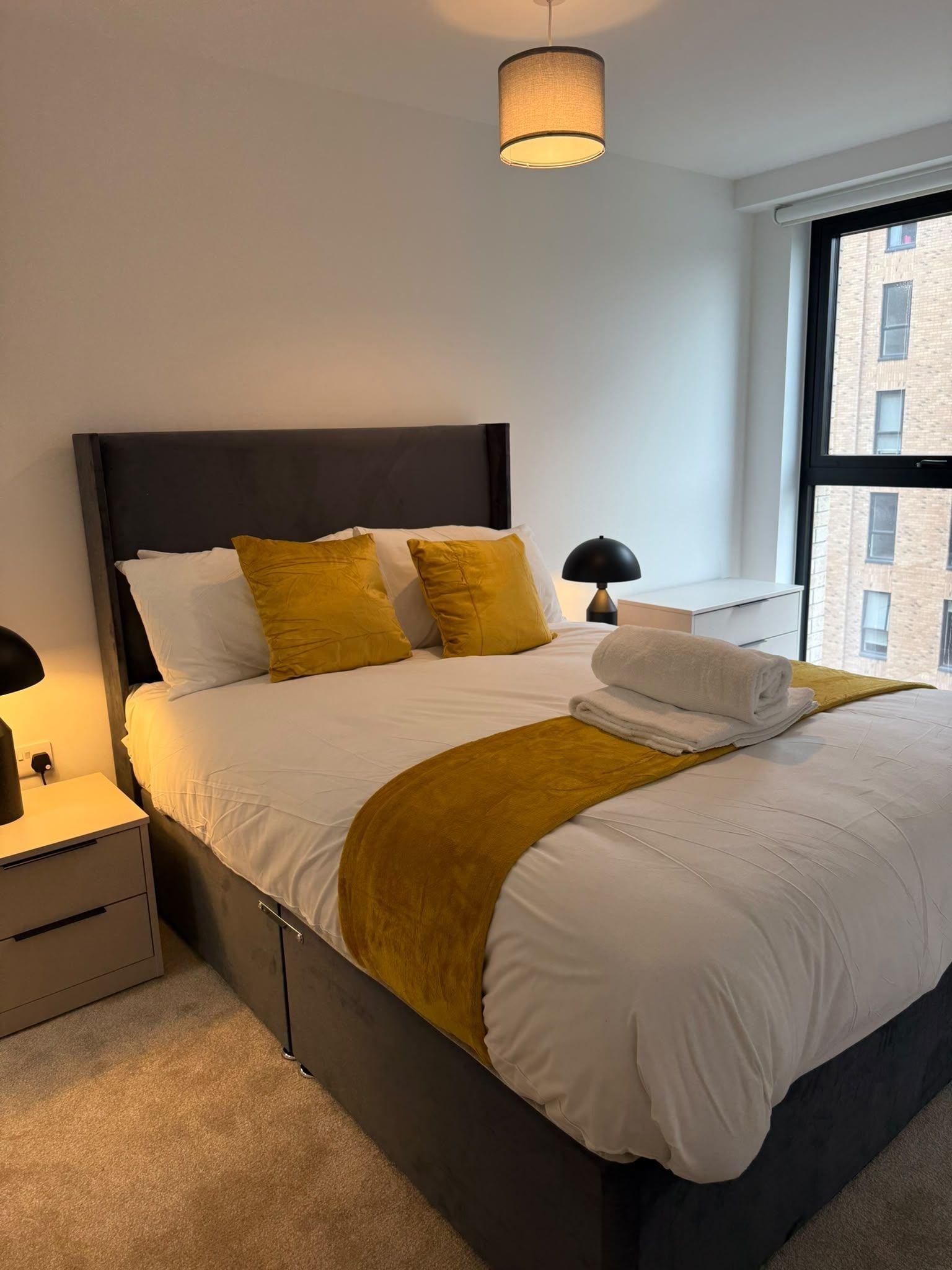 A modern hotel room with a large bed, a bedside table, a lamp, and a window with a view of buildings.