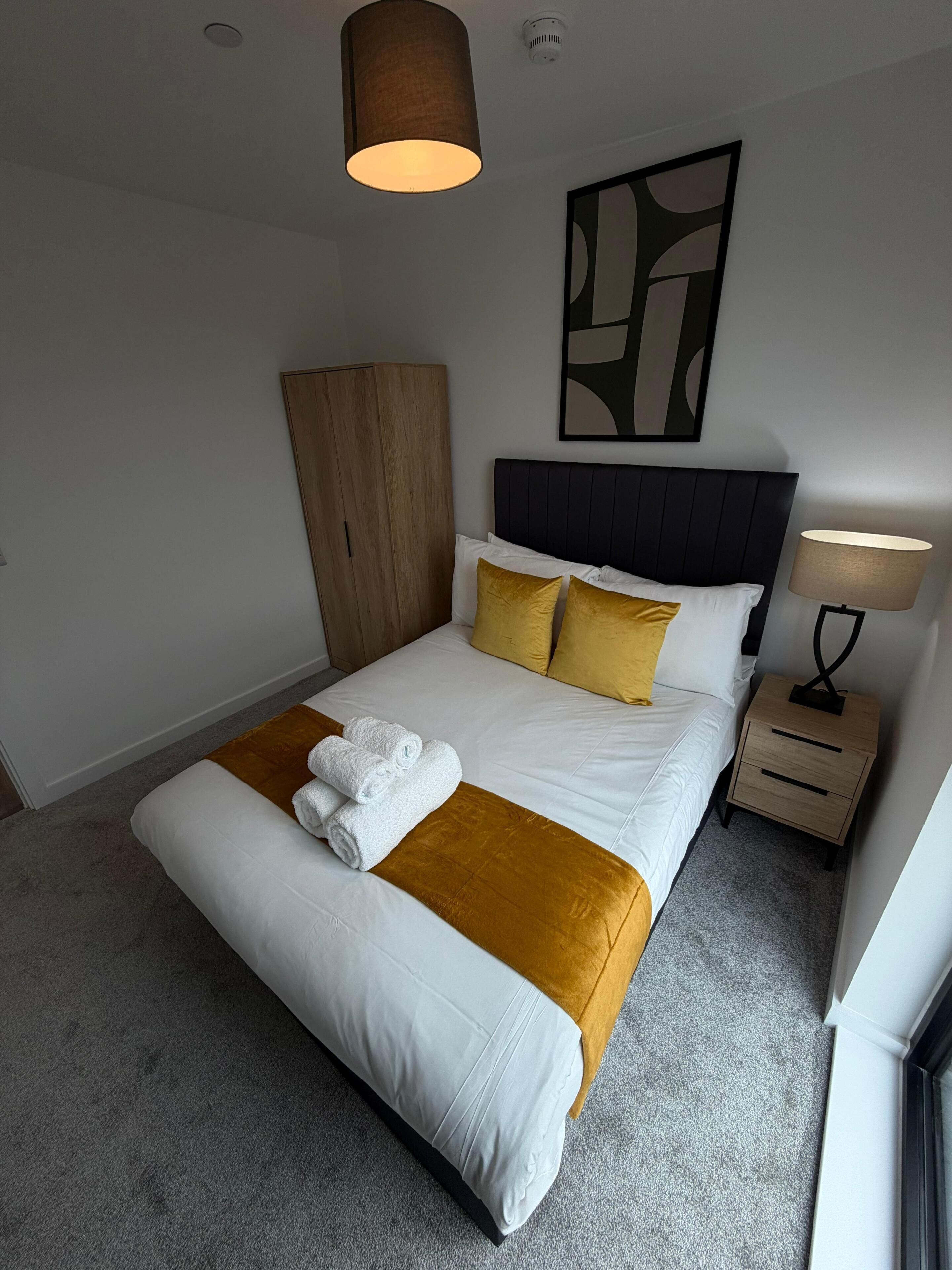 A neatly made bed with white linens and a gold accent, a wooden wardrobe, a modern headboard, a wall-mounted abstract art piece, and a bedside table with a lamp.