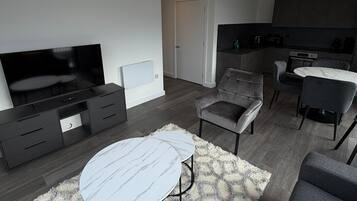 Un salon moderne avec un téléviseur à écran plat, un canapé, une table basse et un coin cuisine à l’arrière-plan.