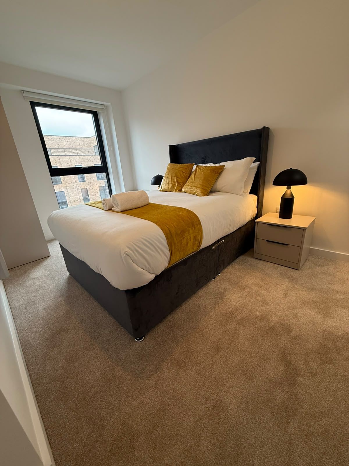 A modern bedroom with a large bed, a nightstand, and a window with a view of buildings.