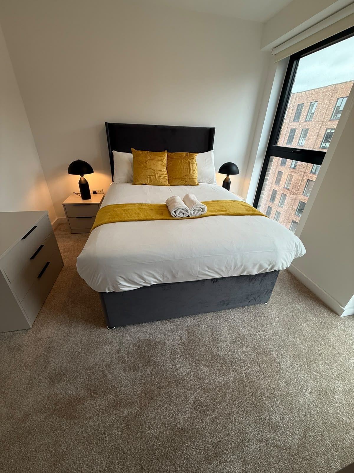Habitación de hotel moderna con una cama grande, dos mesitas de noche, un ventanal con vistas a la ciudad y suelo alfombrado.