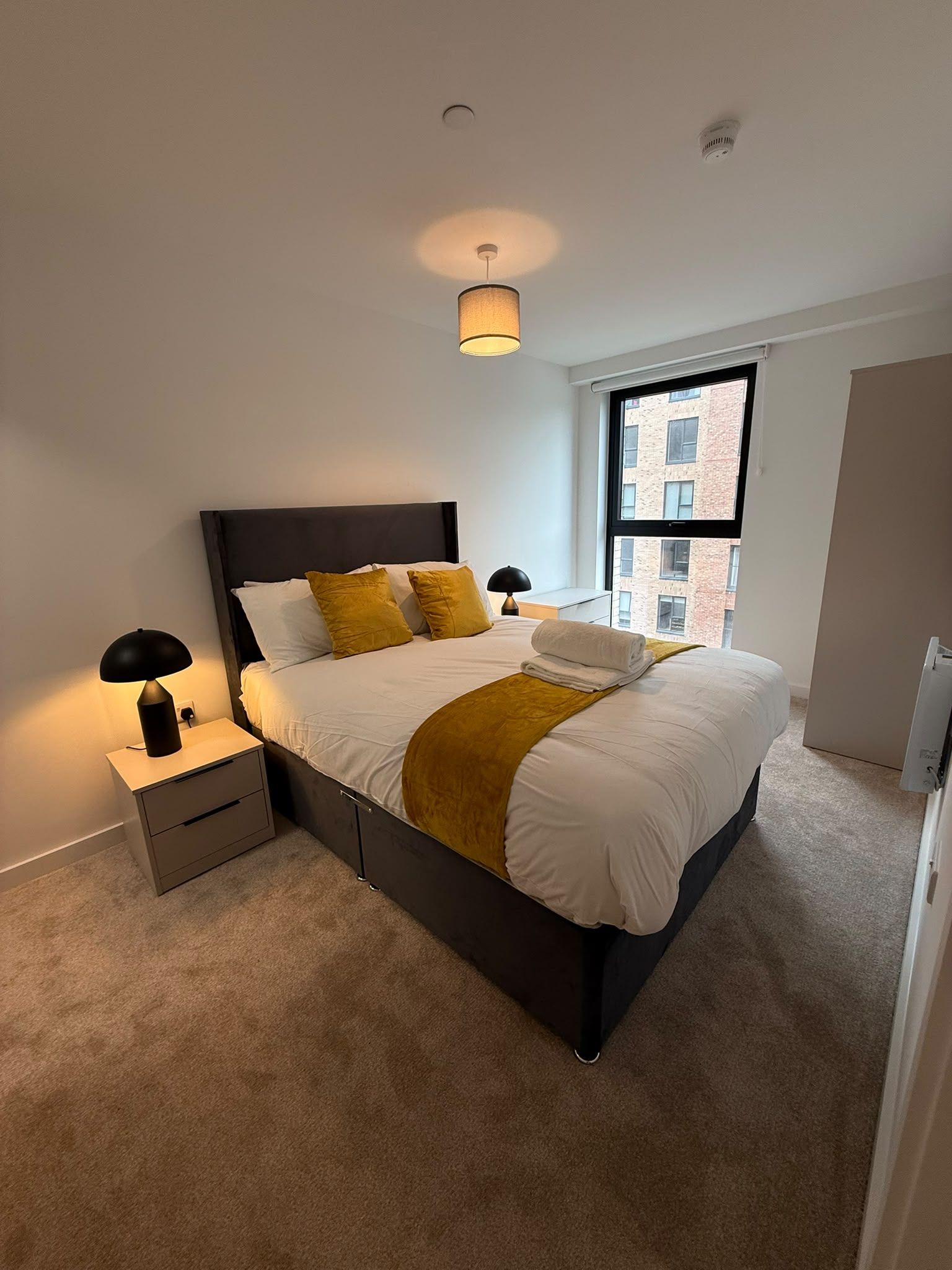 A modern bedroom with a large bed, bedside lamps, a window with a view of buildings, and a ceiling light.