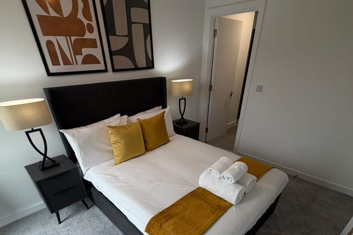 A neatly made bed with white linens, two gold pillows, and a brown throw blanket. There are two bedside tables with lamps, a door to another room, and abstract wall art above the bed.