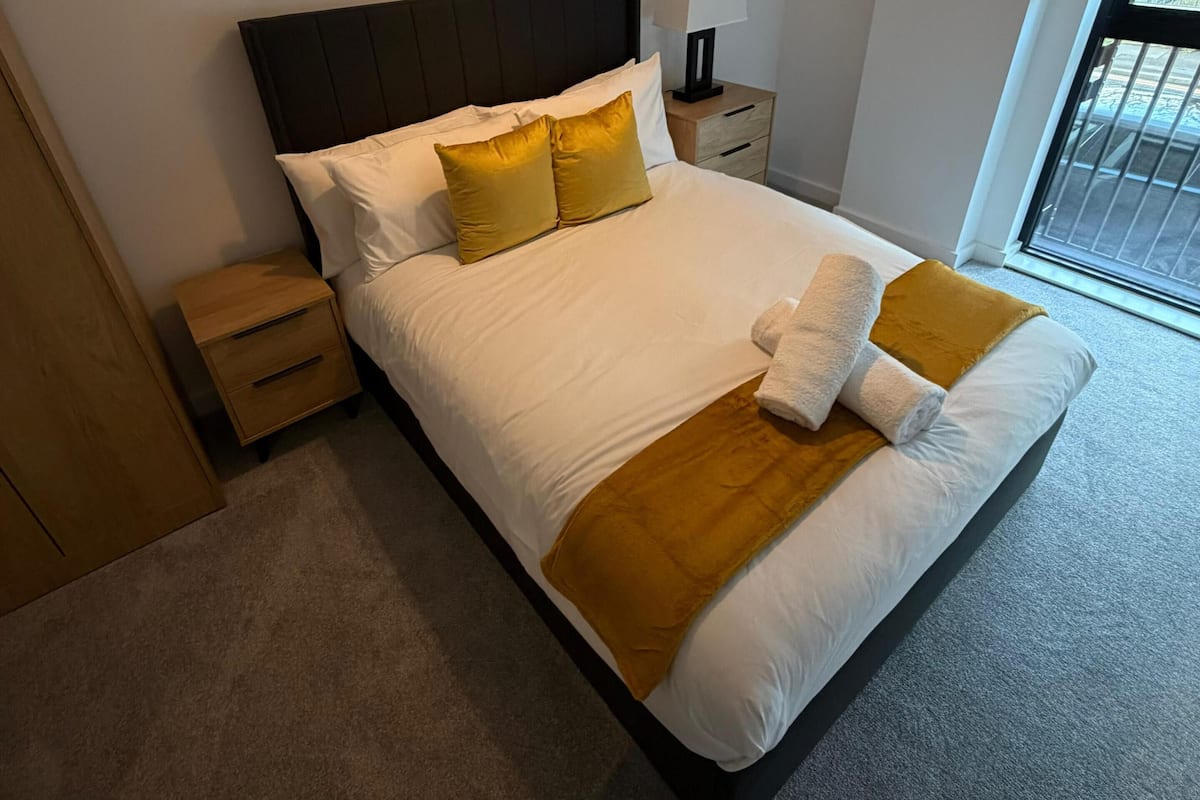 A neatly made bed with white and mustard bedding, a wooden nightstand with a lamp, and abstract wall art above the bed.