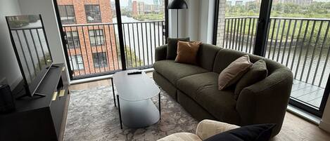 A modern living room with a sofa, armchair, and a coffee table. There is a large window with a view of buildings and a balcony.