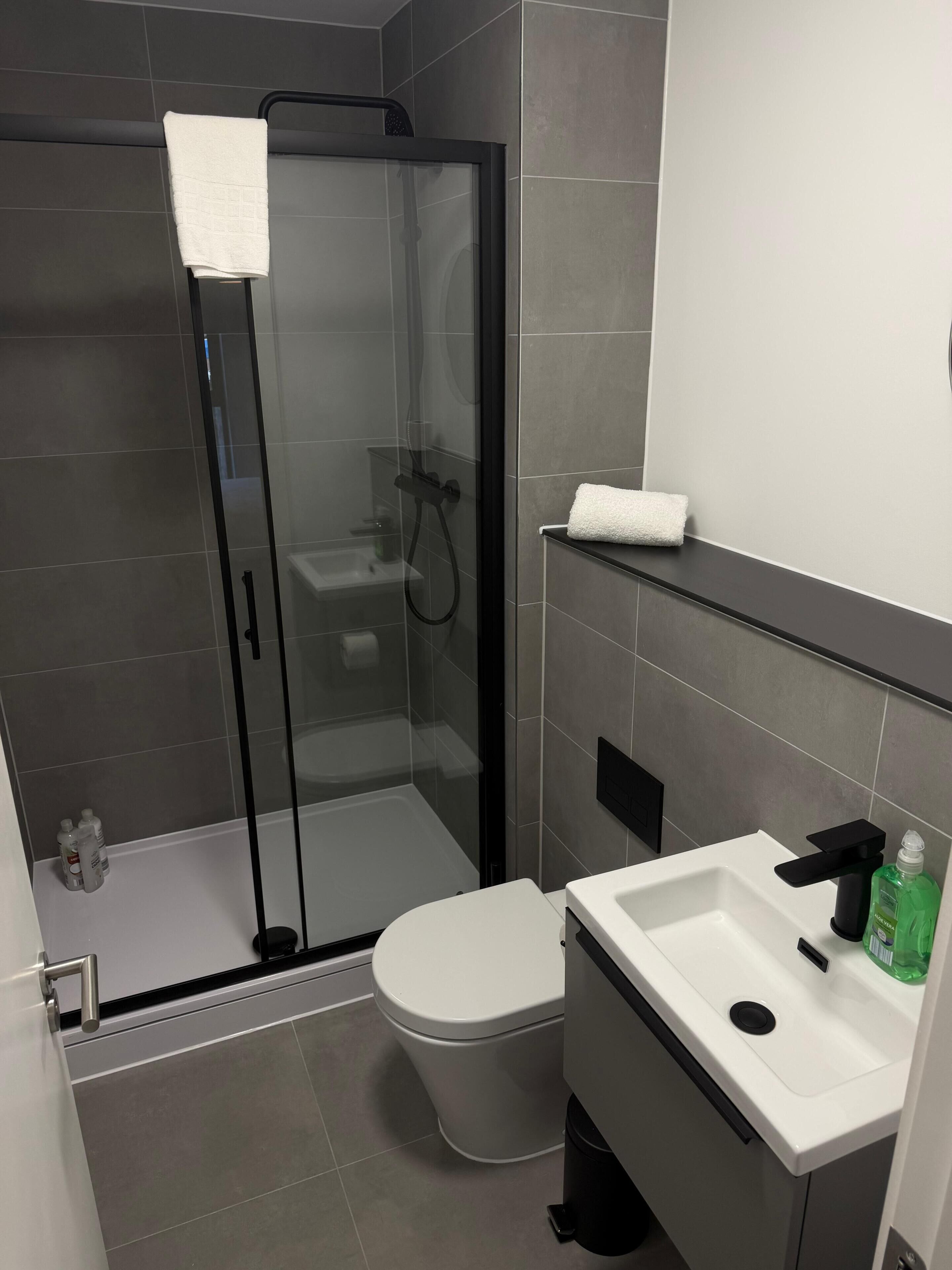 Et moderne badeværelse med en brusekabine i glas, et hvidt toilet og en hvid vask med sort armatur.