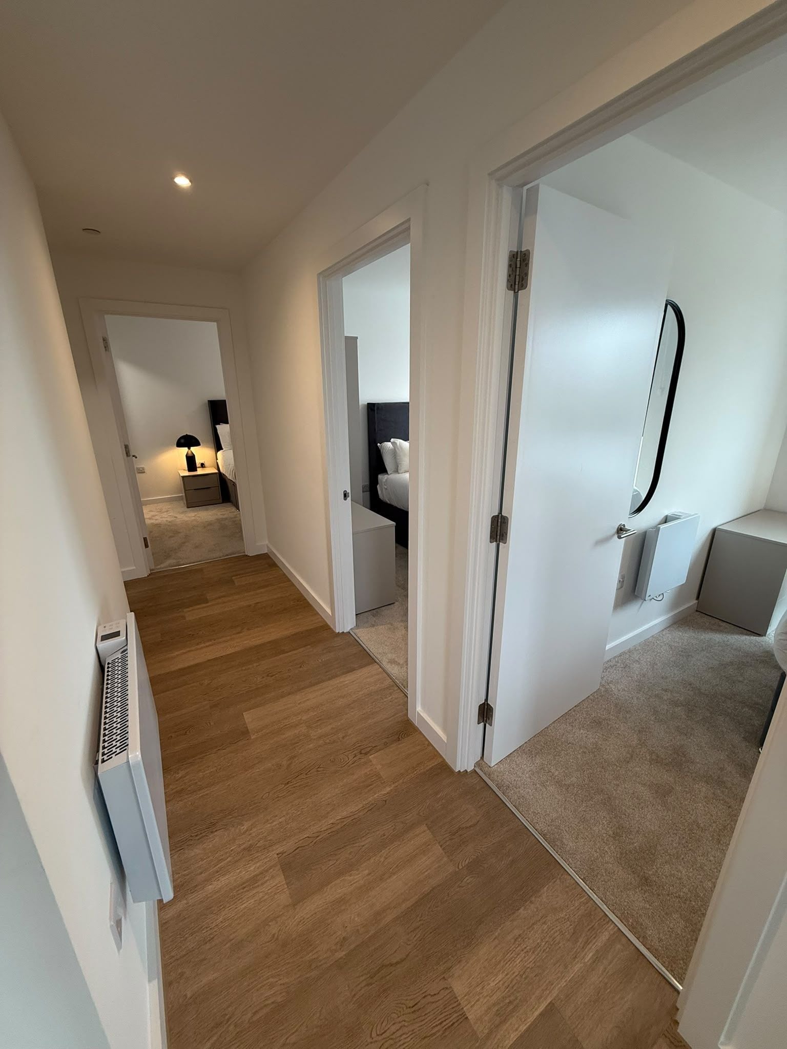 Un couloir au plancher de bois menant à une chambre à coucher équipée d’un lit et d’une table de chevet.