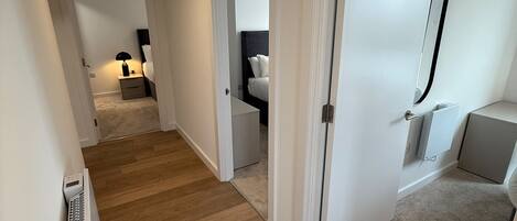 Un couloir au plancher de bois menant à une chambre à coucher équipée d’un lit et d’une table de chevet.