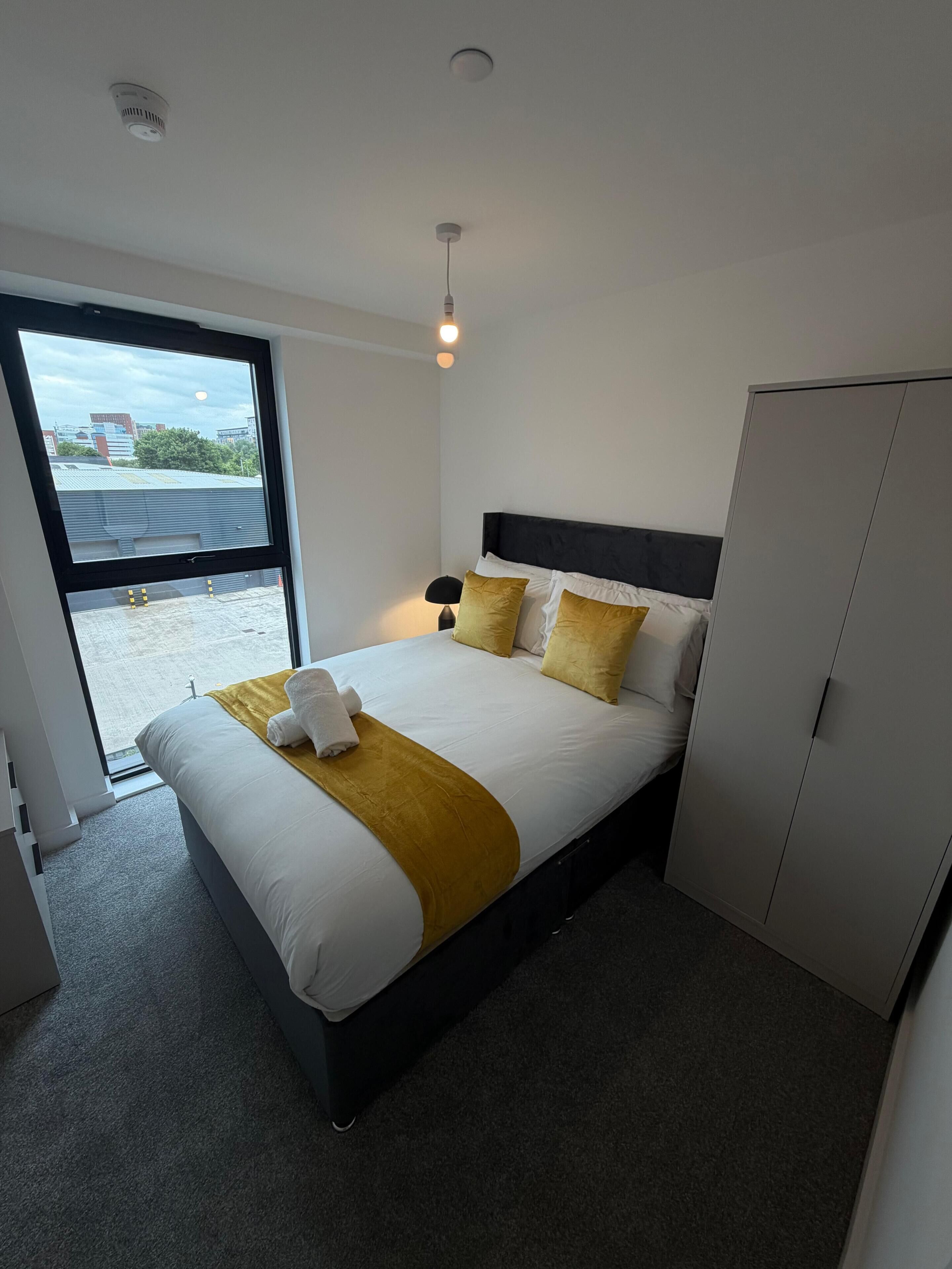 A modern hotel room with a large bed, a wardrobe, a bedside lamp, and a view through a large window.