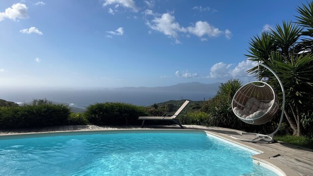 Ein klares, blaues Schwimmbecken mit einer Holzterrasse, einem Hängestuhl und Blick auf das Meer und die Berge.