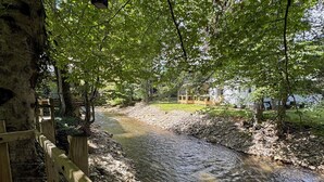 Una terraza de madera con vistas a un río y un puente de piedra, rodeada de árboles y un edificio al fondo.