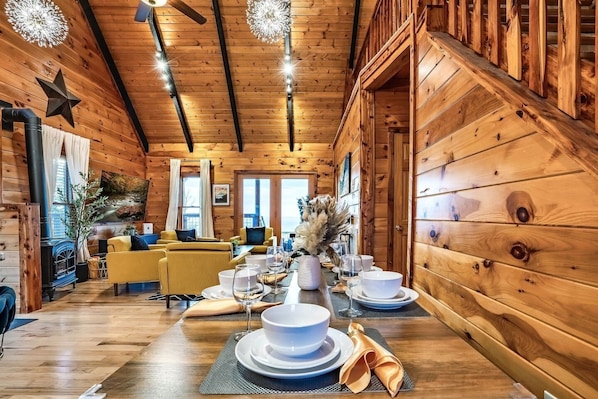 A wooden cabin interior with a dining table set for four, a sofa, and a fireplace. - Amazing Sunset+MountainViews+HotTub•Firepit•Games•Sleeps 8 (Sevierville)