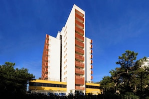 A high-rise building with multiple balconies, a yellow bus in the foreground, and trees in the background. - Seafront Apartment on the Promenade (Lignano Sabbiadoro)