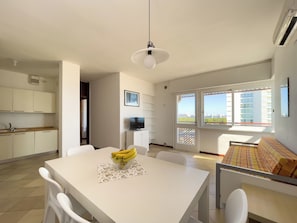 A modern kitchen with white cabinets, a dining table with chairs, a TV, and a balcony with a view of buildings. - Seafront Apartment on the Promenade (Lignano Sabbiadoro)
