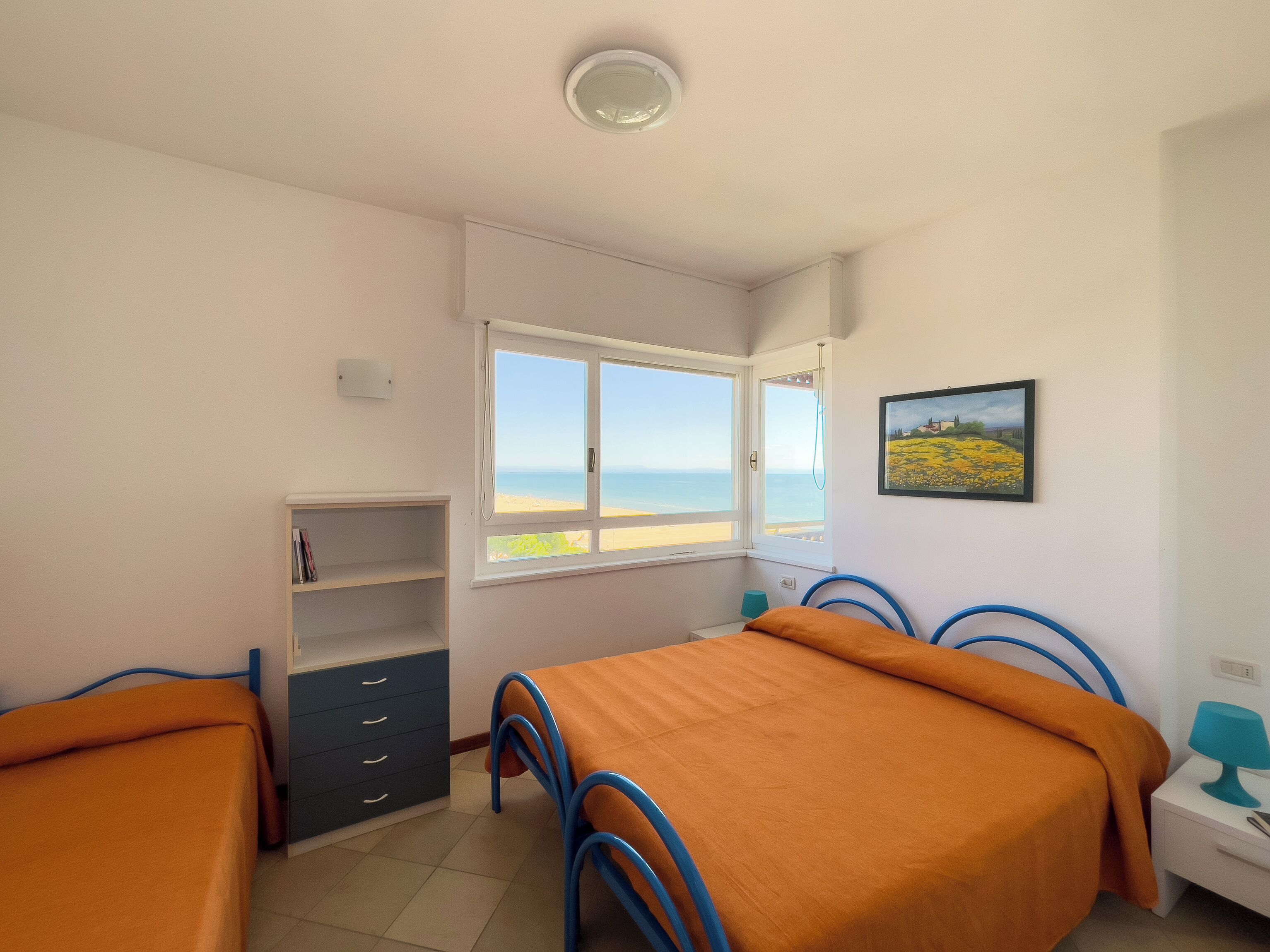 A bedroom with two beds, a window with a view of the sea, a shelf with books, and a painting on the wall.