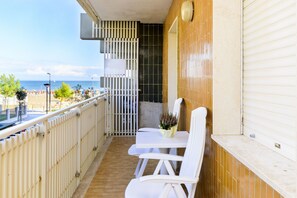 Ein Balkon mit weißen Stühlen und einem Tisch, der Blick auf einen Strand mit Sonnenschirmen und Menschen bietet.