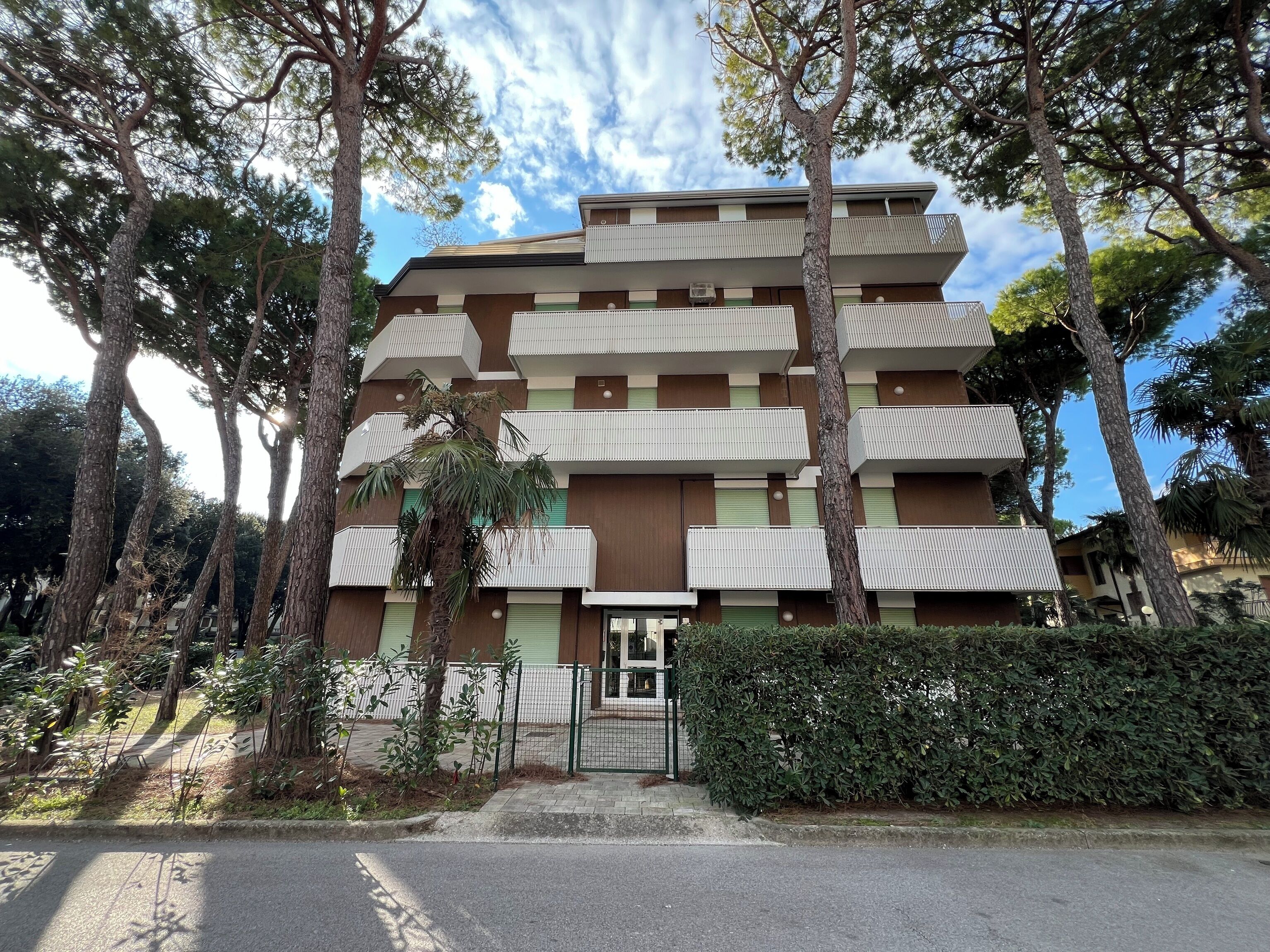 A multi-story building with balconies, surrounded by trees and a fence.