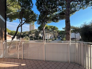 A balcony with a white railing, a tiled floor, and a view of trees and buildings in the background. - Lignano Pineta Flat With Balcony (Lignano Sabbiadoro)