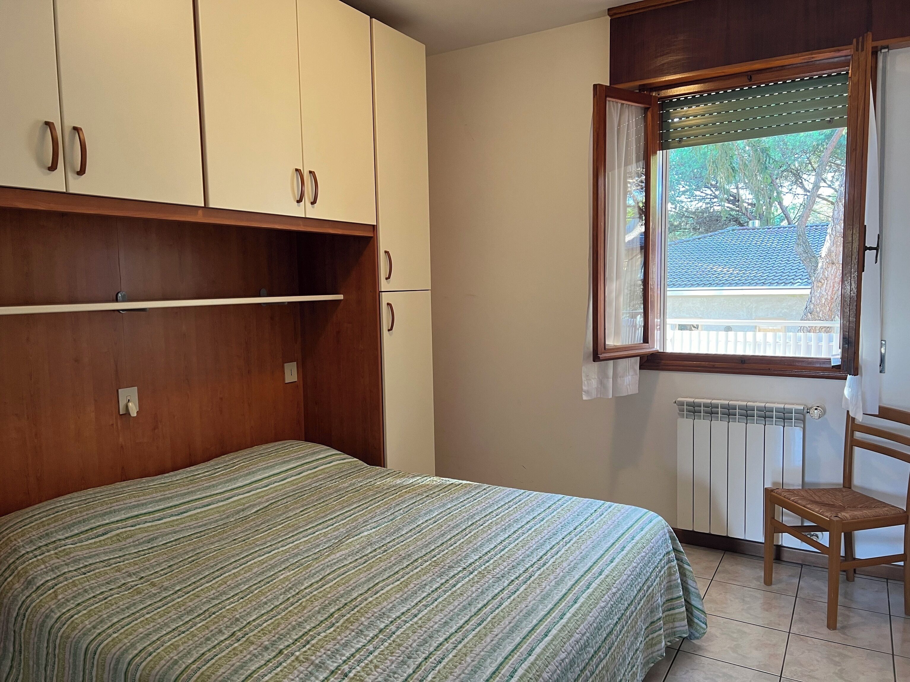 A bedroom with a bed, wooden wardrobe, a chair, a radiator, and a window with a view of the outside.