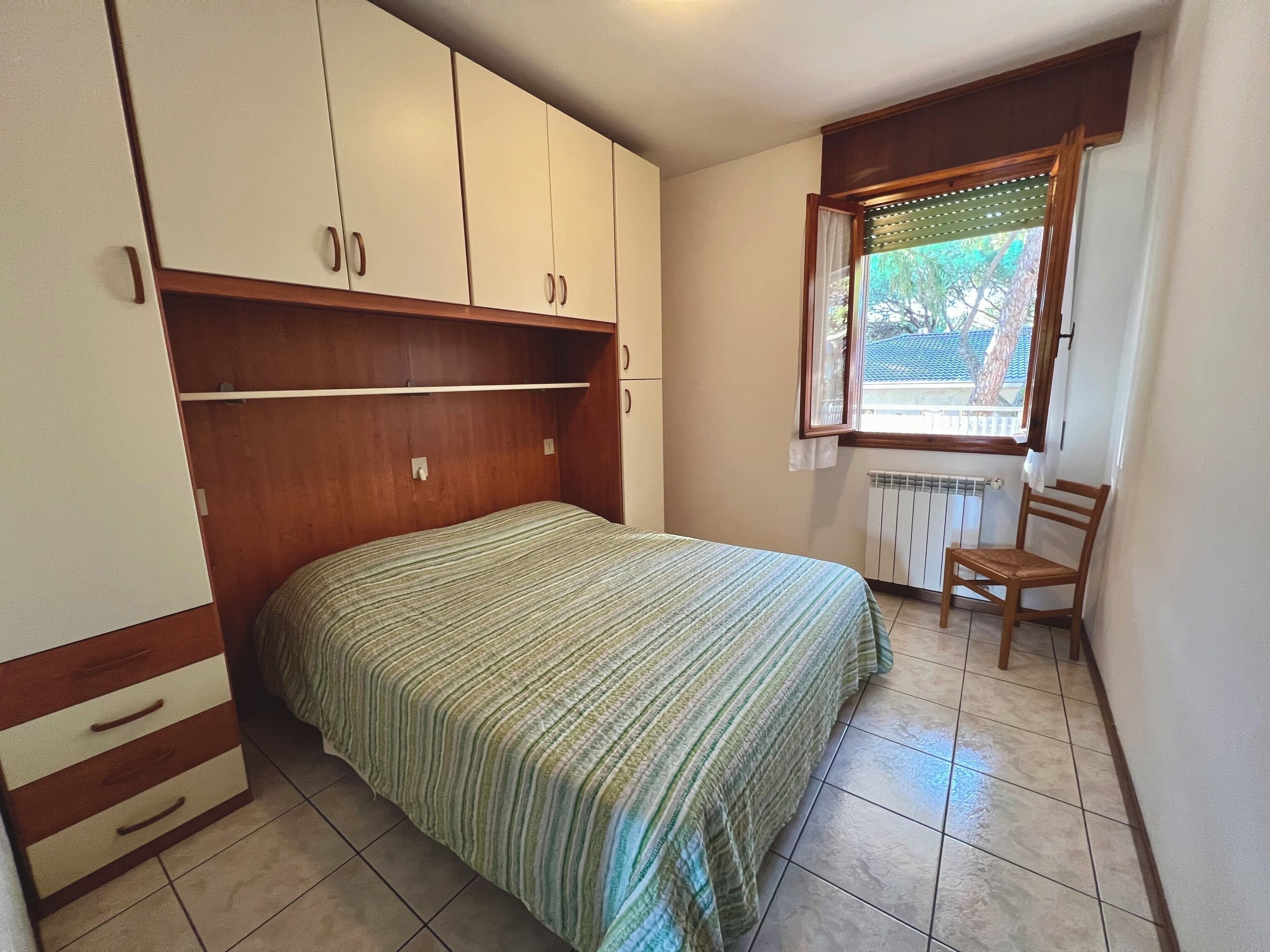 A bedroom with a bed, wooden wardrobe, chair, and a window with a view.