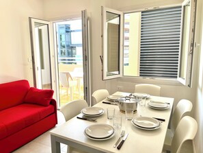 A dining area with a table set for four, a red sofa, and a balcony with a view of buildings. - Flat With Parking Near the Beach (Lignano Sabbiadoro)