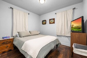 Een slaapkamer met een bed, gordijnen, een dressoir, een televisie en twee ingelijste foto's aan de muur.