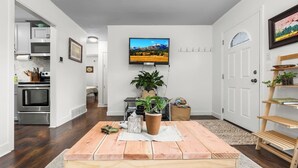 Uma sala moderna com uma mesa de jantar de madeira, uma TV de tela plana na parede, uma cozinha com eletrodomésticos de aço inoxidável e um corredor que leva a outro cômodo.