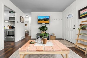 Una sala de estar moderna con una mesa de comedor de madera, un televisor de pantalla plana montado en la pared, una cocina con electrodomésticos de acero inoxidable y un pasillo que lleva a otra habitación.