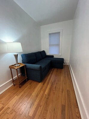 Ein Eckzimmer mit einem dunkelblauen, modularen Sofa, einem kleinen Holztisch, einer Stehlampe und einem Fenster mit Jalousien.