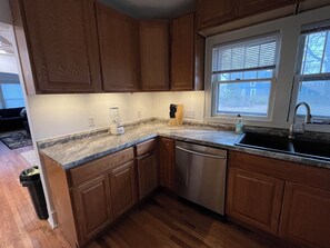 A kitchen with wooden cabinets, a granite countertop, a dishwasher, a window with blinds, and a sink. - Black River Bungalow with 3 bedrooms (Port Huron)
