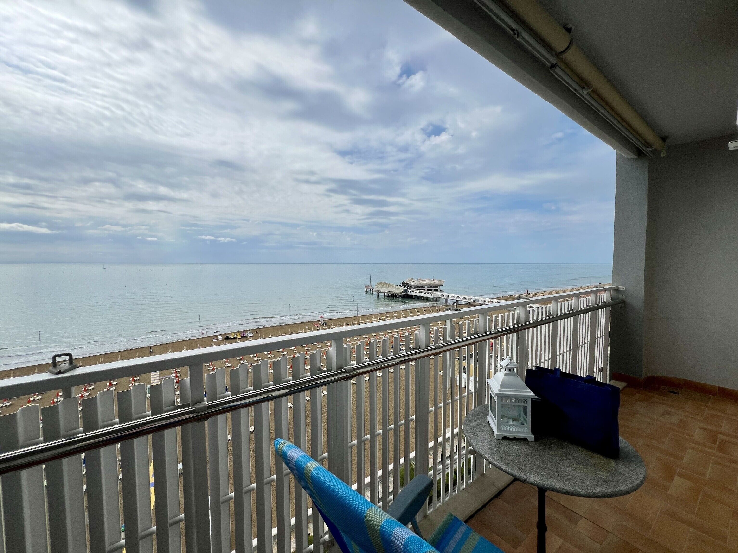 A balcony with a view of the sea, a pier, and a beach with umbrellas.