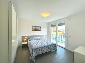 Un dormitorio con cama, mesita de noche, lámpara de pared, luz de techo, ventana con vistas y una puerta.