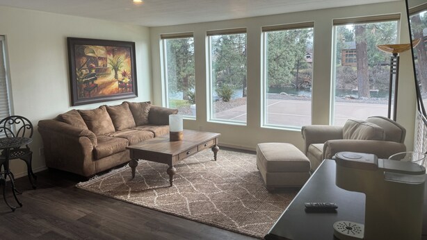 A living room with a sectional sofa, a coffee table, a side table, and a lamp. There are large windows with a view of a park and a building. - WATERFRONT PARADISE (Spokane)