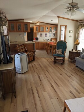 A spacious living room with a wooden floor, a dining area, and a kitchen with wooden cabinets. - CAMP : 2-bedroom mobile home camp - lake access to Chazy Lake in Ellenburg Depot (Ellenburg Depot)