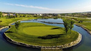 Un campo de golf con un estanque, árboles y casas al fondo.