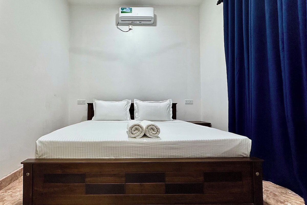 Un dormitorio con cama, almohadas, una toalla enrollada, un equipo de aire acondicionado y una cortina azul.
