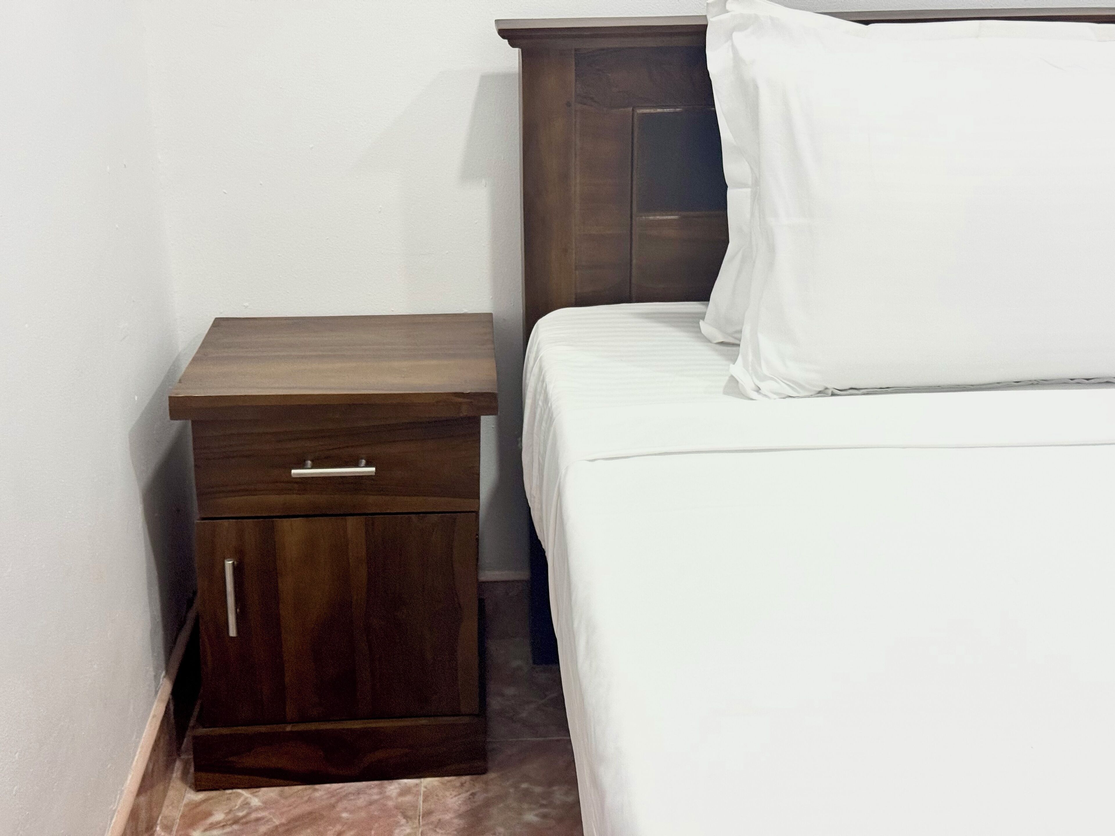 Una mesita de noche pequeña y oscura de madera con un cajón, una cama blanca con un almohadón y una pared lisa.