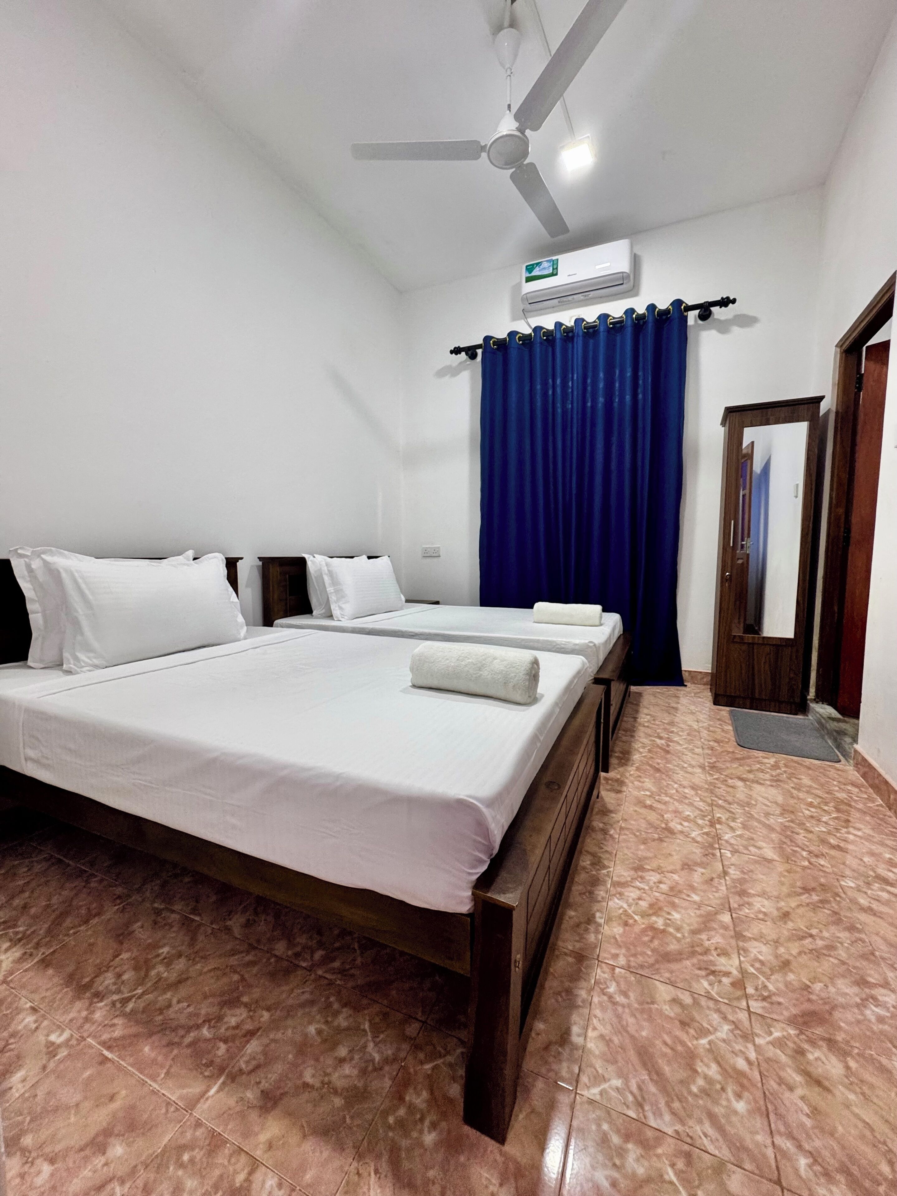 Une chambre d’hôtel avec un lit, un ventilateur de plafond, un appareil de climatisation, un rideau bleu et une porte en bois.