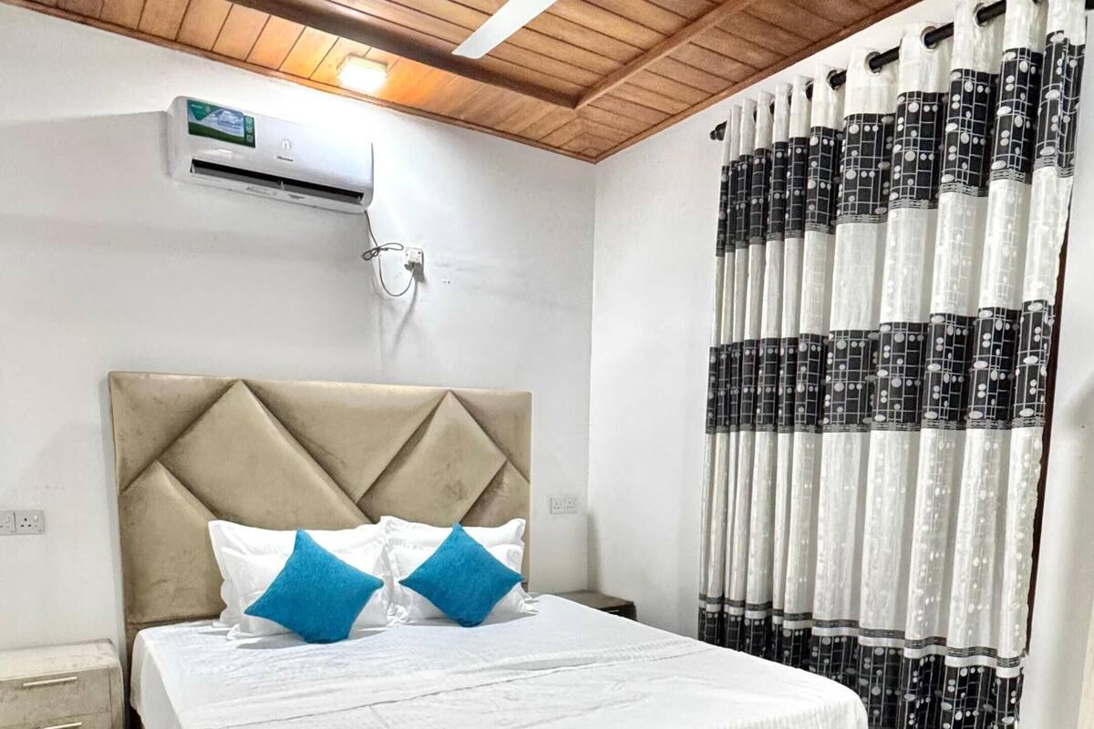 Un dormitorio con cama, ventilador de techo, aire acondicionado, mesita de noche y ventana con cortinas estampadas.