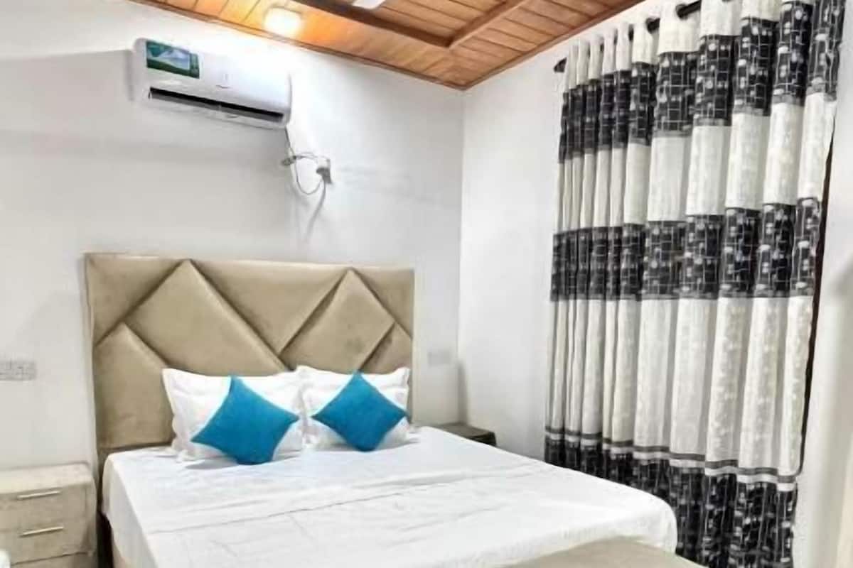 Un dormitorio con cama, ventilador de techo, unidad de aire acondicionado, mesita de noche y ventana con cortinas.