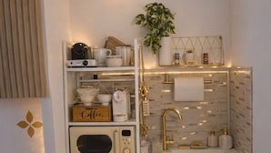 Una kitchenette compacta con cafetera, microondas, refrigerador pequeño, pileta con detalles dorados y una planta en maceta.