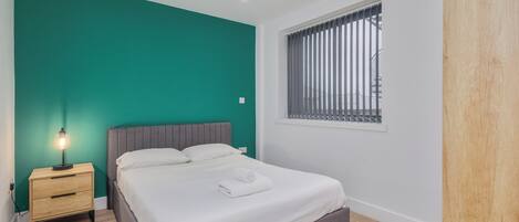 A bedroom with a bed, a nightstand, a lamp, a window with blinds, and a teal wall.